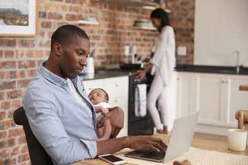 Father On Laptop Holds Newborn Son As Mother Makes Meal