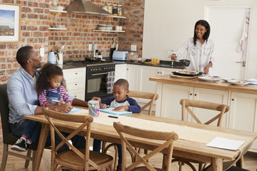 Father And Children Drawing At Table As Mother Prepares Meal