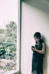 Pretty asian girl holding smart phone and listening to music with her headphones near window