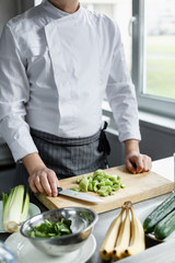 Crop faceless shot of male im uniform cutting celery on cutting board for preparing smoothie.
