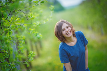 girl in a denim dress laughs, standing between the rows of young green apple trees