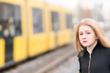 Young blonde girl posing. On background, out of focused yellow streetcar passing
