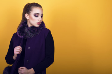 Fototapeta premium Portrait of young gorgeous model with ponytail and artistic make-up wearing trendy purple sleeveless coat with fur collar and looking aside. Isolated on yellow background. Copy-space