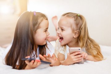 people, children, technology, friends and friendship concept - happy little girls with smartphones lying on floor at home education, school are using a smartphone and smiling while sitting on sofa