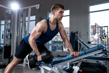 Fototapeta na wymiar Young muscular man in a vest doing one-arm dumbbell rows on bench in gym