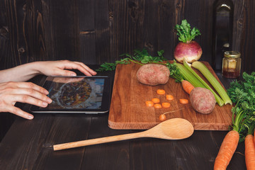 Person looking at digital tablet and cooking healthy food in the kitchen, cutting vegetables on the wooden table