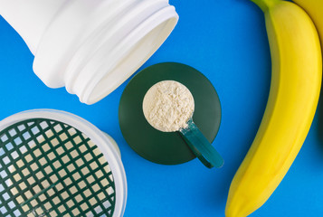 Protein powder in the measuring plastic spoon, shaker bottle with milk and two banana on blue background, sports nutrition after training, top view