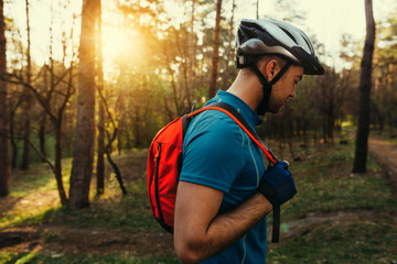 Naklejka premium Portrait in profile of handsome young bearded man cyclist wearing protective helmet, blue t-shirt and red backpack, looking down, thinking about route and cycling. Travel concept. Sport lifestyle.