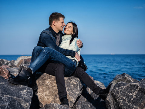 Emotional Couple Sitting On The Rocks On The Beach With Ocean On Background. She Is Sitting On His Lap And Smiling