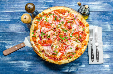 Round pizza with meat, tomatoes and herbs, cutlery and spices, on a blue denim background