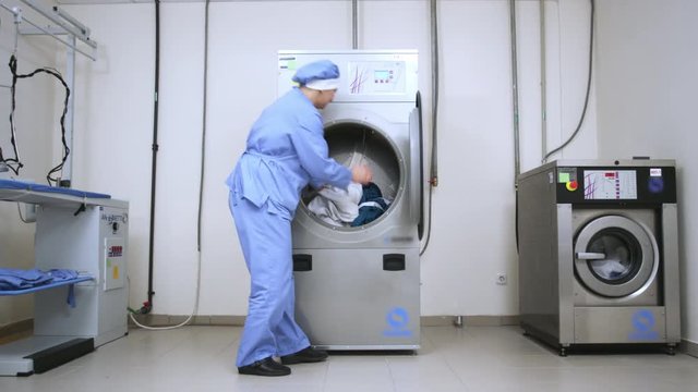 Female worker take away clothes from drying machine at laundry room. Woman removing cloths from washing machine at industrial laundry. Dryer laundry machine in laundry service
