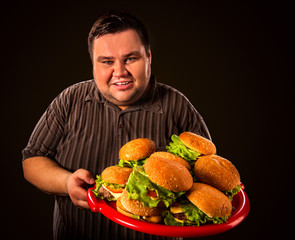 Fat man eating fast food hamberger and carries treat for friends on tray. Breakfast for overweight person. Junk meal leads to obesity. Person regularly overeats concept on black background.