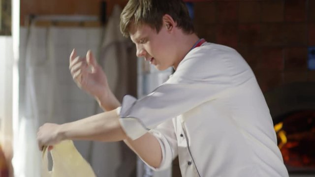 Slow motion of young man spinning and tossing pizza dough up and then poking finger through it, looking at camera and smiling