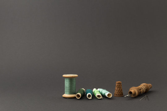 Different rolls with green polyester and silk thread, needles, and a wooden carved needle container with grey background
