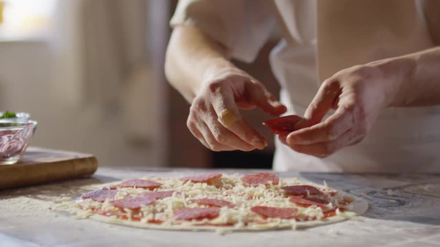 Tilt up slow motion shot of young chef putting salami slices on pizza base 