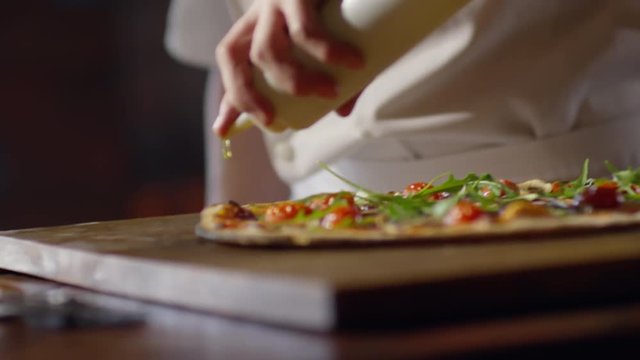 Slow motion shot of young chef in uniform pouring olive oil on top of freshly baked pizza 
