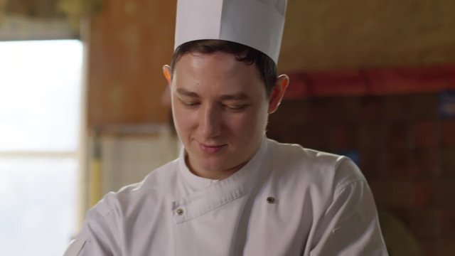 Slow motion of young man in chef uniform and white toque on his head sifting flour while cooking in restaurant kitchen