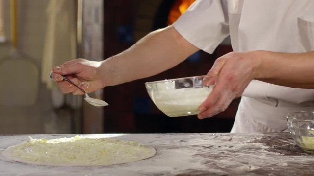Tracking slow motion shot of chef of pouring white pizza sauce with table spoon on dough with shredded cheese on it
