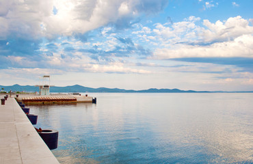 Fototapeta na wymiar Stone quay and a watchtower near the sea shore against the background of a hill range and dramatic cloudy morning sky. Zadar, Croatia