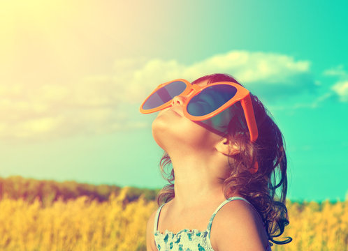 Happy little girl with big sunglasses looking at the sun in the wheat field in summer
