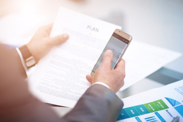 Texting to colleague. Confident young man in suit holding smart phone and looking at it while sitting at his working place