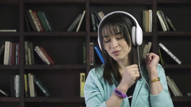 Young brunette woman in headphones is listening to music and dancing against book shelves
