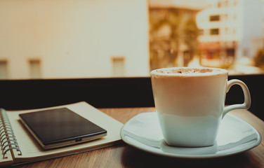 Capuccino coffee,notebook and mobile on wooden table the windows background