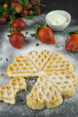 Eine frisch gebackenen Herzwaffel mit Puderzucker und frischen Erdbeeren und einem Schälchen mit Sahne