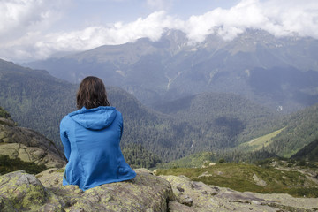 Woman in blue sweater sitting on the edge of a cliff and looking on a beautiful mountain landscape, illuminated by the sun