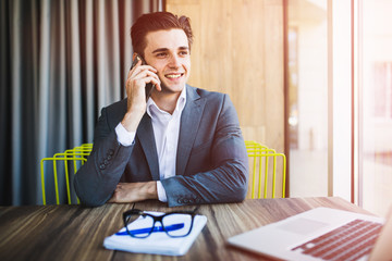 Portrait of handsome young male in glasses sitting at office desk with laptop computer and talking on mobile phone.