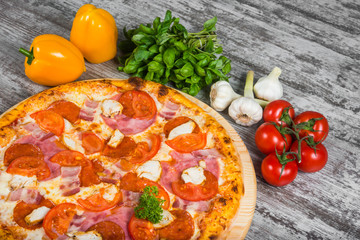 Pizza with sausage, chicken, bacon, ham, with rosemary and spices on a light wooden background. Italian pizza on a background of green basil and fresh vegetables