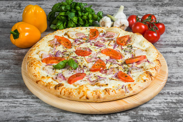 Meat pizza with bacon, chicken, tomatoes, with rosemary and spices on a light wooden background. Italian pizza on a background of green basil and fresh vegetables
