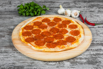 Thin pizza chilli with sausage salami (pepperoni) with tomato sauce, rosemary and spices on a light wooden background. Italian pizza on a background of green basil and fresh vegetables