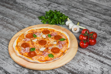 Thin pizza with salami (pepperoni), bacon and chicken with tomato sauce, rosemary and spices on a light wooden background. Italian pizza on a background of green basil and fresh vegetables