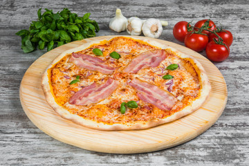 Thin garlic pizza with bacon, tomatoes and onions. Italian pizza on a background of green basil and fresh vegetables