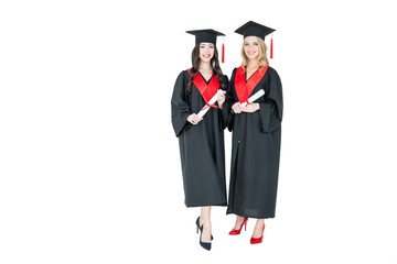 Two beautiful young students in academic caps holding diplomas and smiling at camera