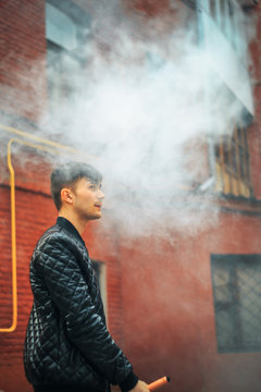 Vape. A handsome young white guy blows steam from an electronic cigarette in a vintage old red yard. Vaping.
