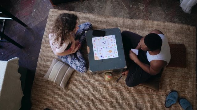 Multiethnic couple in pajamas sitting on the floor and playing the board game. Man and woman laugh and show the monkey.