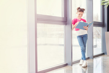 Fototapeta na wymiar Full length view of smiling little girl standing near window and reading book