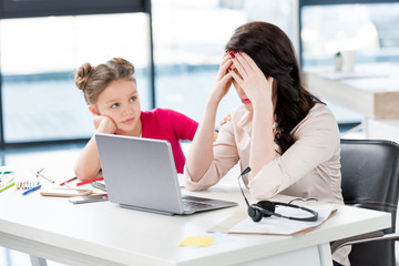 Little girl looking at stressed mother working with laptop in office