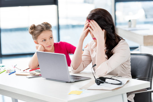 Little Girl Looking At Stressed Mother Working With Laptop In Office