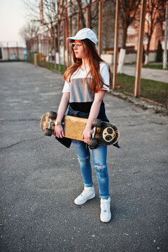 Young teenage urban girl with skateboard, wear on glasses, cap and ripped jeans at the yard sports ground.