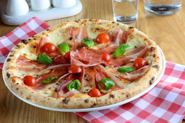 Delicious pizza with prosciutto, mozzarella, cherry tomatoes and basil, served on a wooden table for a dinner in italian restaurant. Italy food background. Pizza from wooden oven.
