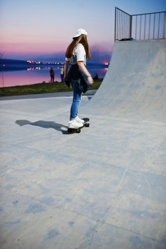 Young teenage urban girl with skateboard, wear on glasses, cap and ripped jeans at skate park on the evening.