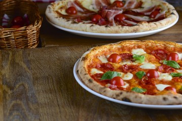 Two hot tasty pizza with pepperoni, prosciutto, mozzarella, cherry tomatoes and basil, served on a dark wooden table for a dinner in a  restaurant. Italy food concept.