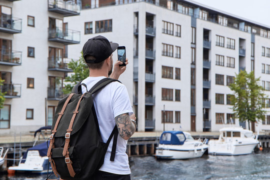 Young hipster man with backpack, taking picture of street on smart phone at journey in Copenhagen, city tourism