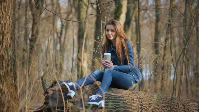 Young girl in jeans clothes in spring park drinks coffee out of paper cup sitting on a log