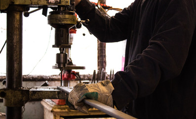 Construction worker drills hole in rod of metal iron using industrial drill in a bright workshop / workshed / factory wearing protective gear and gloves