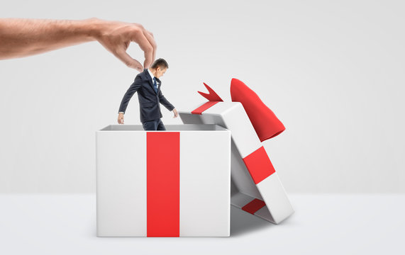 A large male hand getting a tiny businessman out from a white gift box with a red bow on white background.
