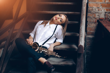 Young attractive girl sitting on steps in white shirt with a saxophone - outdoor. Sexy young woman...